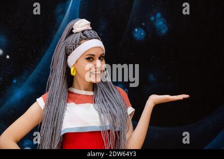 Portrait of excited young woman with dreadlocks or kanekalon pigtails looking at camera and smiling pointing with hand somewhere wearing white headban Stock Photo