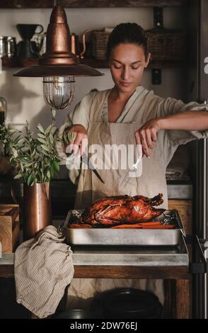 Young woman in apron carving whole oven roasted duck for Thanksgiving holiday or Christmas party celebration in kitchen interior. Traditional Autumn o Stock Photo