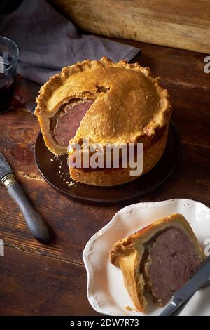 Traditional british raised pork pie with bacon and herbs with one slice cut and served next to a glass of red wine Stock Photo
