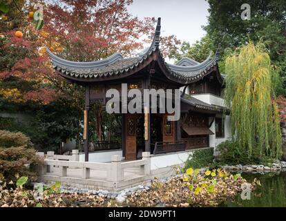 A pavilion or building in the Lan Su Garden in Portland, Oregon with a lake in the foreground and trees surrounding the wooden building. Stock Photo