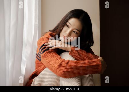 Depressed sad girl feeling lonely sitting by home winter looking out the window in cold weather winter. Asian woman feeling unhappy. Concept of depression, loneliness, anxiety, grief emotions Stock Photo