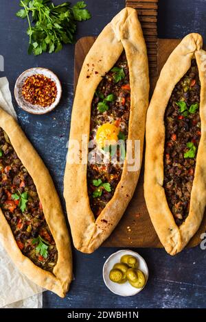 Turkish pide stuffed with beef and an egg on a wooden board and on the ground accompanied by crushed chillies, pickles and parsley photographed on a d Stock Photo
