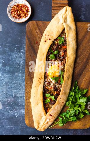Turkish pide with beef and egg topping on a wooden board accompanied by parsley and red pepper flakes on a dark background. Stock Photo