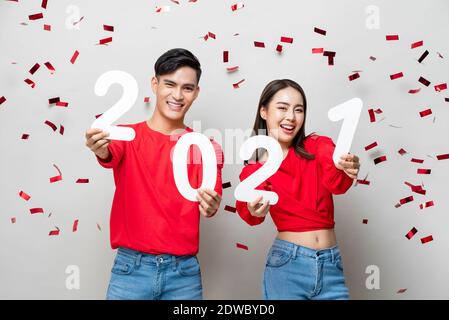 Smiling happy Asian couple in red casual attire holding 2021 numbers for new year concept on light gray background with confetti Stock Photo