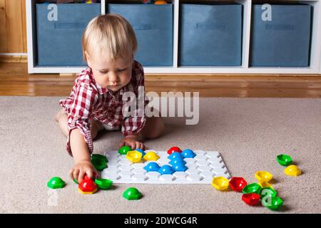 A toddler plays with a plastic mosaic on the carpet in the children's room. Early development, the Montessori method. Stock Photo