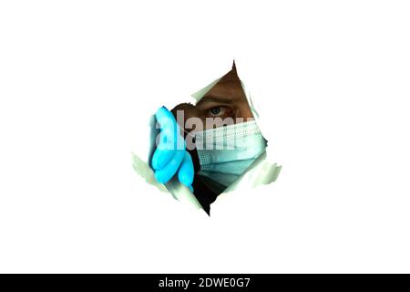 A man in a mask and gloves looks through the hole. Situation a man in a medical mask peeps through a hole in the wall Stock Photo