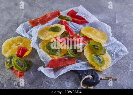 Dried fruits: Yellow candied pineapple rings, red papaya and green on white wrapping paper on gray concrete Stock Photo