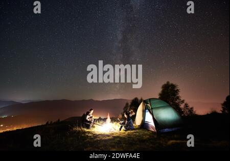 Young happy couple travellers having a rest at bonfire beside camp and illuminated tourist tent under night sky full of stars and Milky way. On the background starry sky, mountains and luminous city Stock Photo