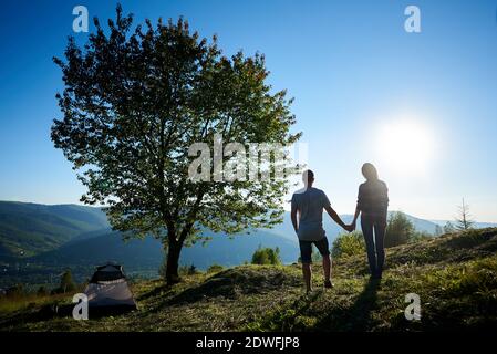 Back view of two young tourists resting near summer camping and big tree in the mountains at sunny morning. Guy and girl holding hands. Tourism adventure active lifestyle concept Stock Photo