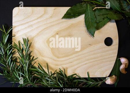 Cutting board of light wood framed with bay leaves, rosemary sprigs and cloves of garlic, designer template Stock Photo