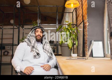 Thoughtful. Arabian businessman working in office, business centre using device, gadget. Modern saudi lifestyle. Man in traditional wear and scarf looks confident, busy, handsome. Ethnicity, finance. Stock Photo