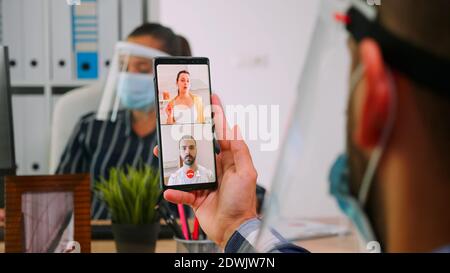 Businessman leader leads distant communication using smartphone for videomeeting talking with remotely employee sitting in new normal office corporate building. Freelancer respecting social distance Stock Photo