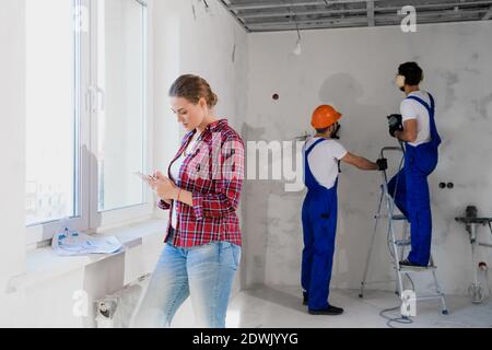 A woman in a plaid shirt is texting by phone. Two construction workers in helmets and headphones are drilling the wall Stock Photo