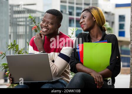 two happy students study online holding multicolor notebooks and a laptop and using it together outdoors Stock Photo