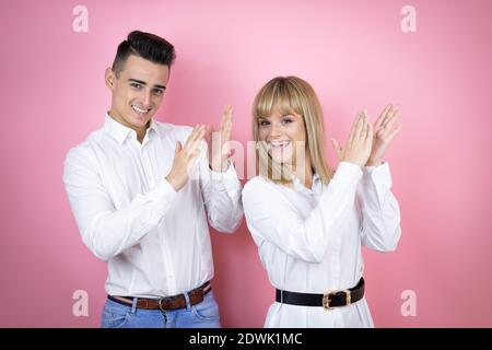 Young couple of girlfriend and boyfriend over isolated pink background clapping and applauding happy and joyful, smiling proud hands together Stock Photo