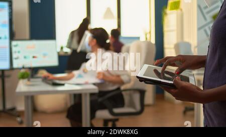 African employee using tablet standing in office room and team of financial analysts working in background. Multiethnic coworkers respecting social distance in business company during global pandemic Stock Photo