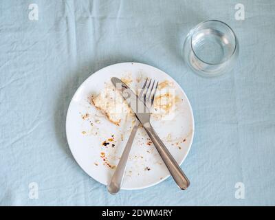 dirty plate after eating on the dining table. Top view of dirty white plate with crosswise cutlery over blue linen tablecloth Stock Photo