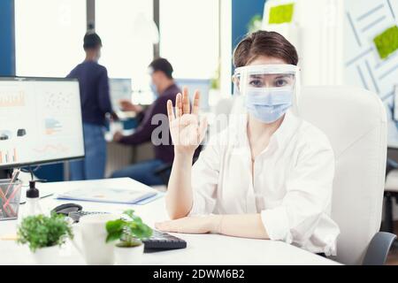Executive wearing face mask and protection shield communicating with client during covid-19..0 Pov of manager speaking with remotely team during online conference while colleagues working in background. Stock Photo