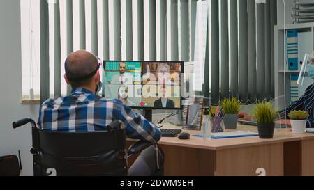Paralysed manager in wheelchair talking with mask and visor during videocall having online conference in business office. Immobilized freelancer working in financial company respecting social distance Stock Photo
