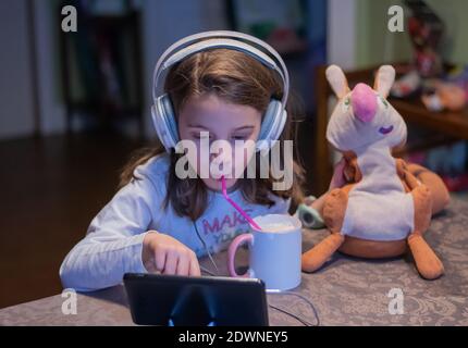 The picture shows a portrait of a little Caucasian girl looking at a tablet while drinking a mug of milk Stock Photo