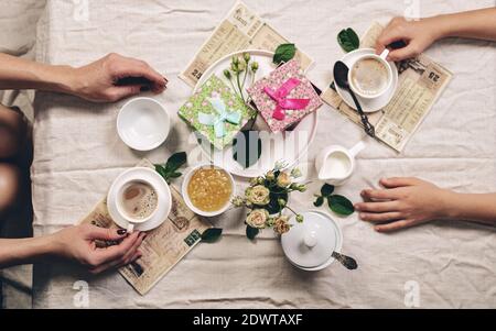 Food still life. White coffee cups, a milk jug with cream, roses and two gifts on a linen tablecloth and graceful female hands. Refined toning. Top vi