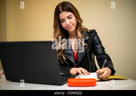 Young woman sitting in front of her laptop, she is doing her homework. She has a smiling expression and is looking at her laptop. Stock Photo