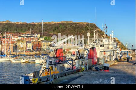 A boat moored in a harbour with a the light from a low sun falling on one side.  A marina and a waterfront are in the background  lying under a hill t Stock Photo