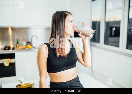 Woman in sportswear drinking sweet banana chocolate protein powder milkshake smoothie.Drinking protein after at home workout.Whey, banana and low fat Stock Photo