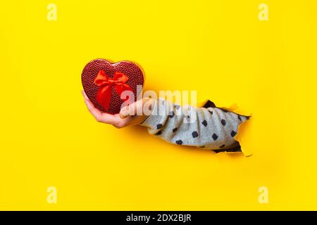 Female hand holds a red heart-shaped gift box on a torn yellow background. Stock Photo