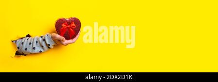 Female hand holds a red heart-shaped gift box on a torn yellow background. Banner Stock Photo