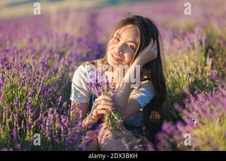 Beautiful young girl with a bouquet of flowers sits in a lavender field in the sunlight Stock Photo
