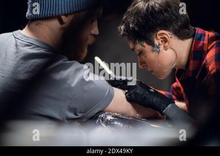 A young girl tattoo artist makes a bull tattoo on the arm of a young hipster man using the handpoke method in a tattoo parlor Stock Photo