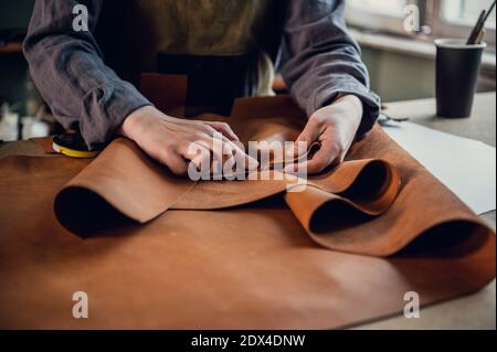 A young apprentice in a boot workshop prepares leather for further use on a large table. Stock Photo