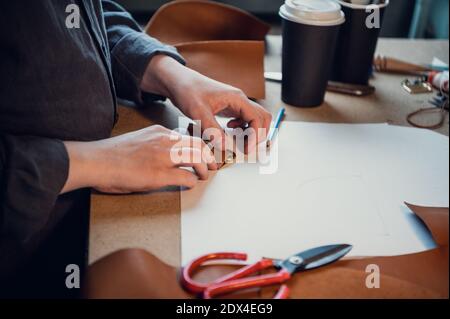 Tailor's hands make loops for the fastener of a women's bag made of genuine leather. Workbench in the workshop. Stock Photo