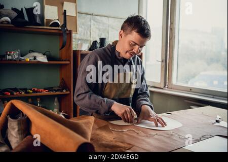 A young apprentice transfers a pattern from paper to leather using a pencil in the workshop on the table. Stock Photo