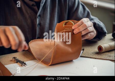 Sitting at a table in the workshop a young guy manually sews leather elements to each other. Stock Photo