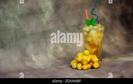 A tall glass of light nectar with ice and a bunch of white grapes nearby on a gray abstract background. Close-up. Stock Photo