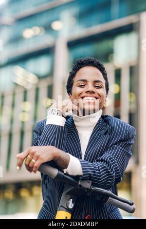 Smiling young businesswoman leaning on electric push scooter Stock Photo