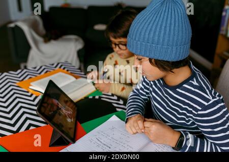 Boy talking to teacher during e-Learning class while male friend studying beside in living room Stock Photo