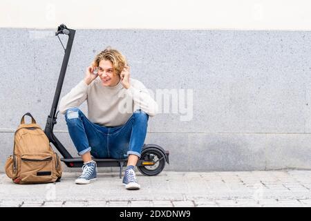 Smiling man with backpack wearing headphones while sitting on electric push scooter against wall Stock Photo