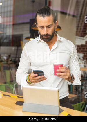 Candid portrait of bearded businessman drinking coffee and using phone inside cafe Stock Photo