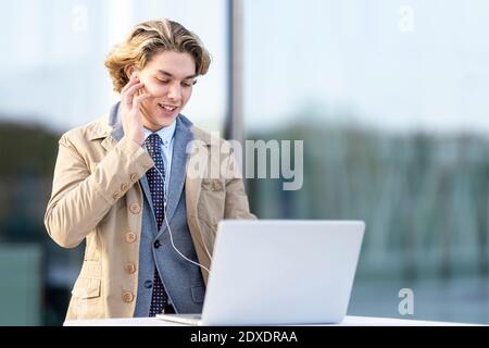 Businessman adjusting in-ear headphones while working on laptop outdoors Stock Photo