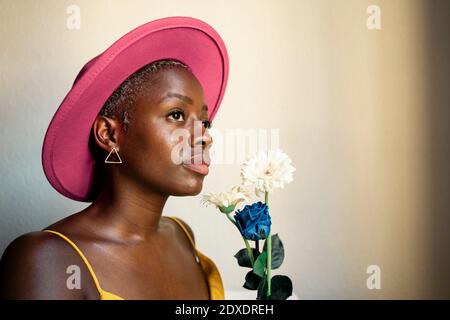 Thoughtful woman wearing pink hat holding flowers while looking away at home Stock Photo