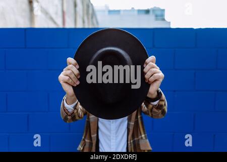Stylish man wearing checked jacket holding black hat in front of face against blue wall Stock Photo