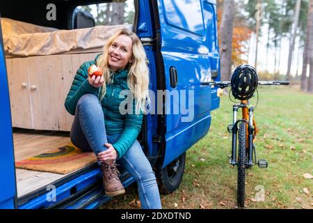 Smiling woman having apple while sitting at door of camper van at Cannock Chase Stock Photo