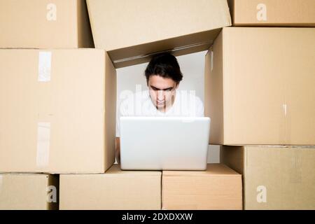 Young man working on laptop under stack of cardboard boxes Stock Photo