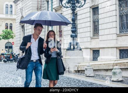 Young businessman and businesswoman walking down the street under an umbrella, texting. Stock Photo