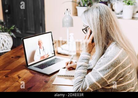 Senior woman talking on mobile phone while writing notes on video call with doctor at home Stock Photo