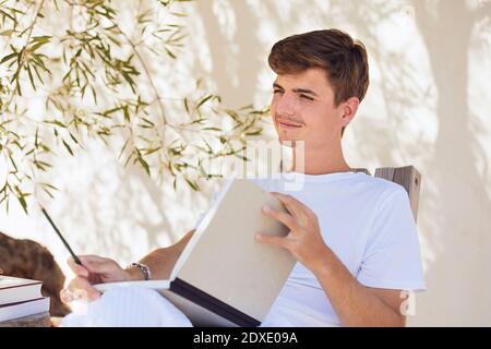 Thoughtful young man with book sitting against wall in backyard Stock Photo