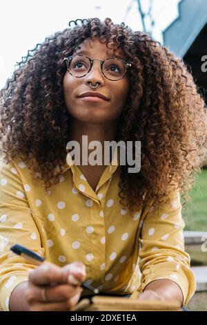 Young thoughtful woman with book looking away while sitting outdoors Stock Photo